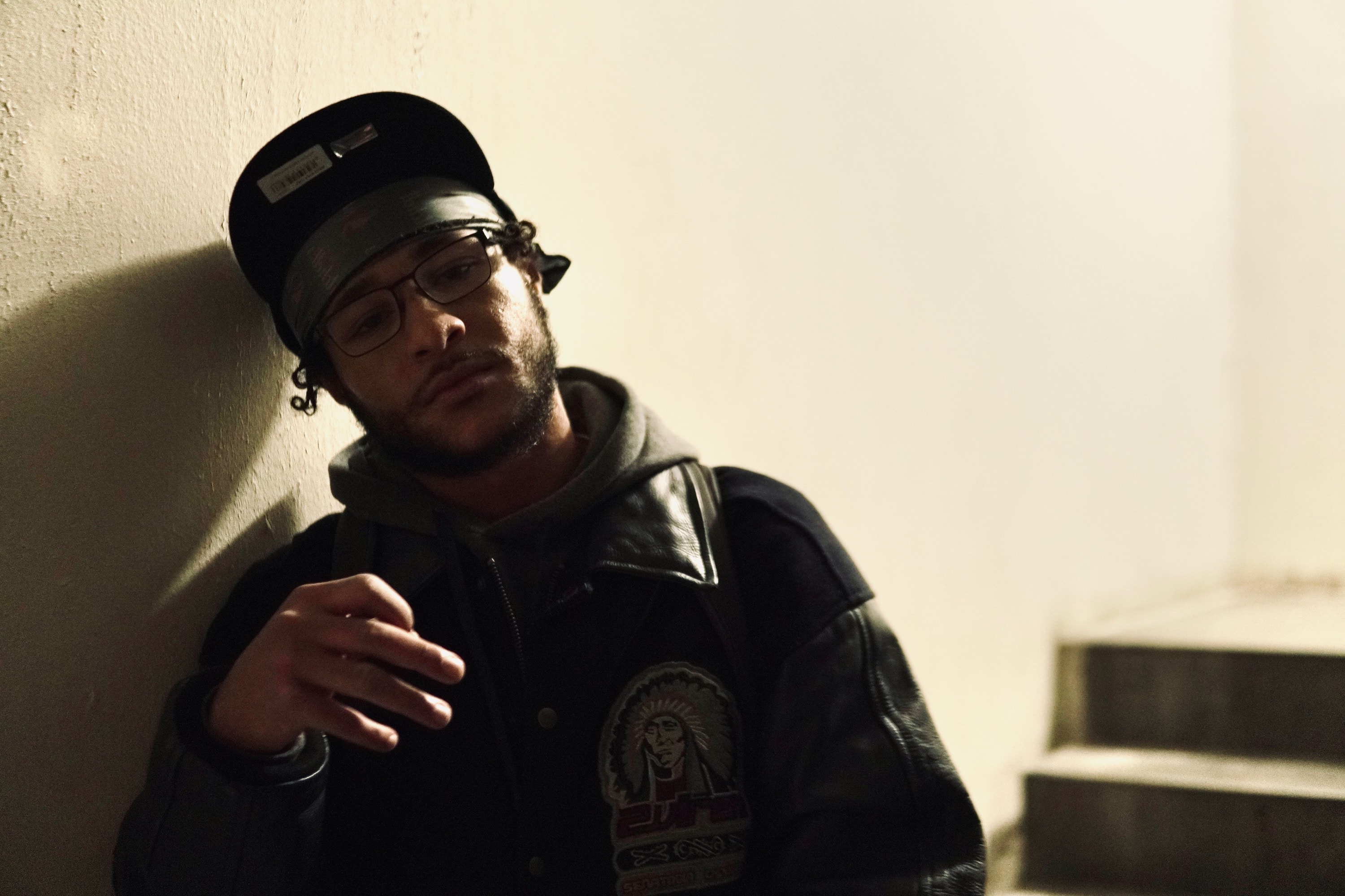 Squintz wearing a cap and glasses leaning against a wall